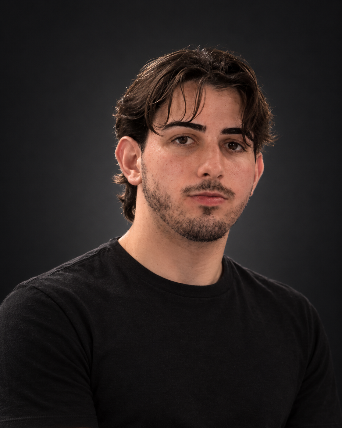 Portrait of Kaio Vinícios in a dark studio, wearing a black T-shirt, with dark hair and a short beard.